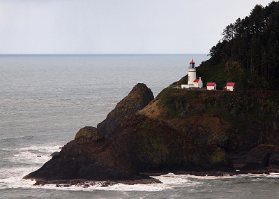 [A white lighthouse with a red cap with several red-roofed white buildings near it. All are perched at the edge of a rock outcropping extending into the ocean. This view was taken from a high vantage point across a small bay.]